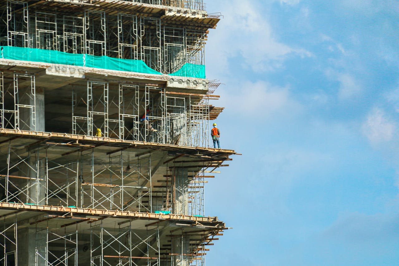 A construction worker stands on a scaffolding at a high-rise building under a blue sky.